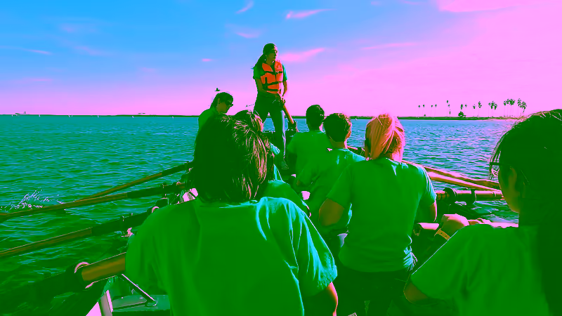A crew rowing a pulling boat out on the harbor as the coxswain steers